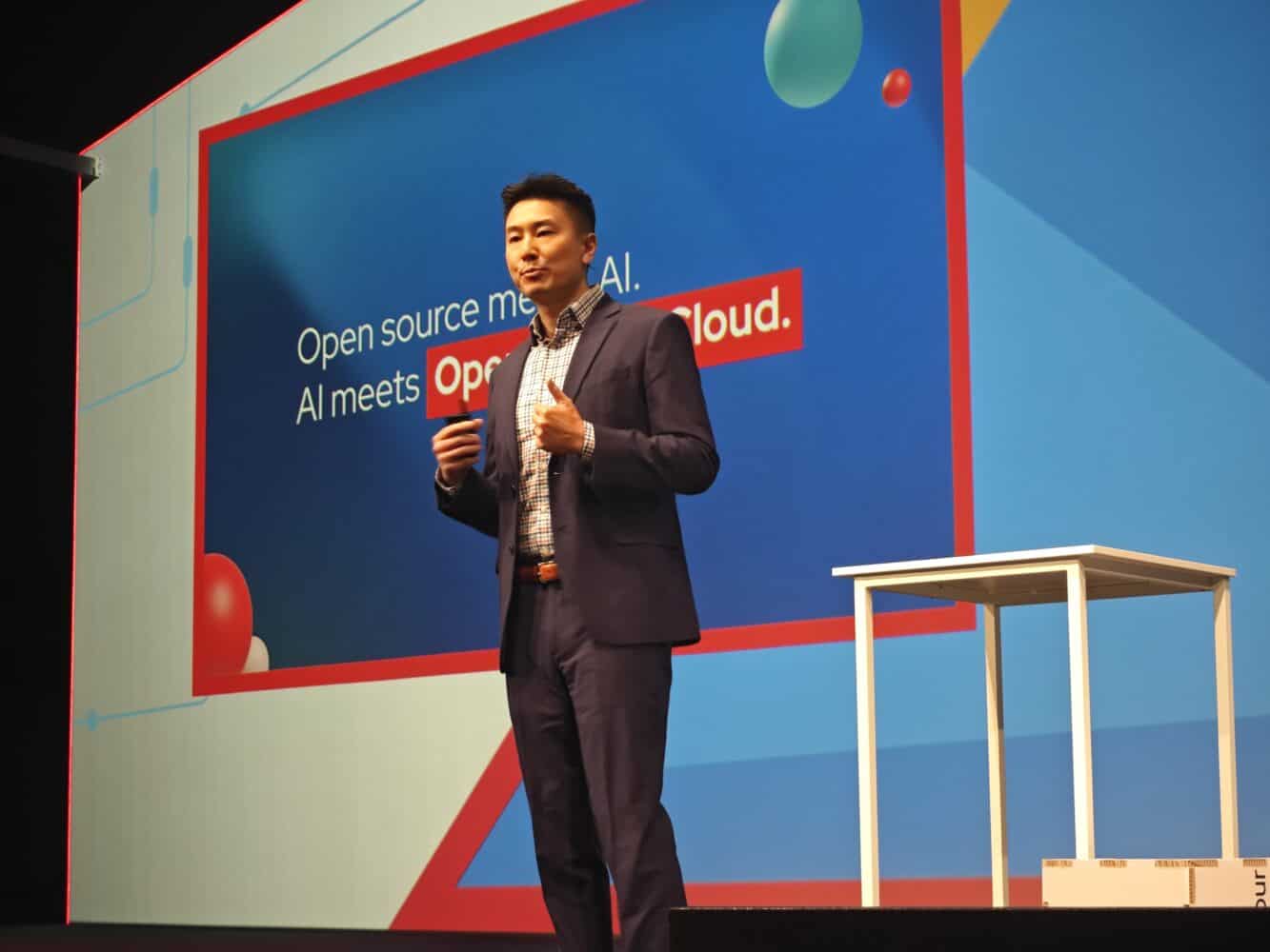 1733997813064 Een persoon in pak spreekt op een podium voor een scherm met daarop een tekst over open source, AI en cloudtechnologie.