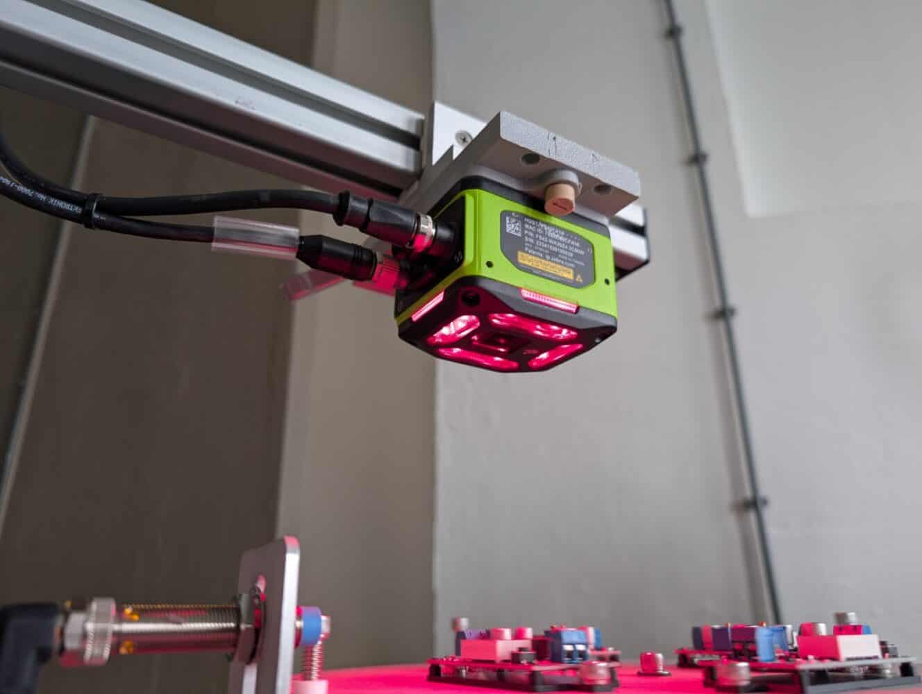 Close-up van een groene industriële camera met rode lampjes, gemonteerd op een metalen frame, gepositioneerd boven elektronische printplaten.