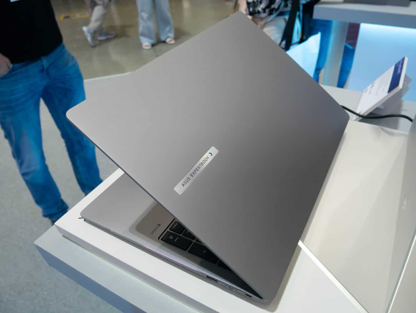 Een zilveren laptop, half opengeklapt op een presentatietafel, met een sticker met het opschrift "ASUS Zenbook". Op de achtergrond staan mensen.